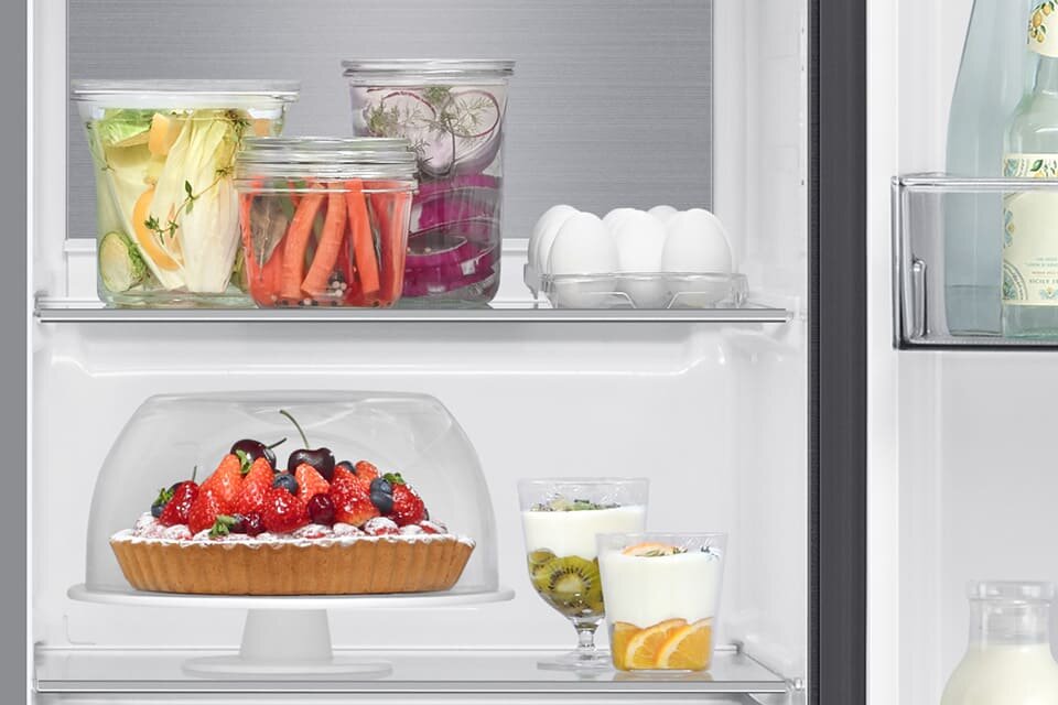 Wnętrze lodówki z półkami, na których znajdują się różne produkty spożywcze, takie jak warzywa w słoikach, jajka, ciasto z owocami pod kloszem oraz jogurty z owocami. Na drzwiach lodówki widoczna jest butelka z napojem.