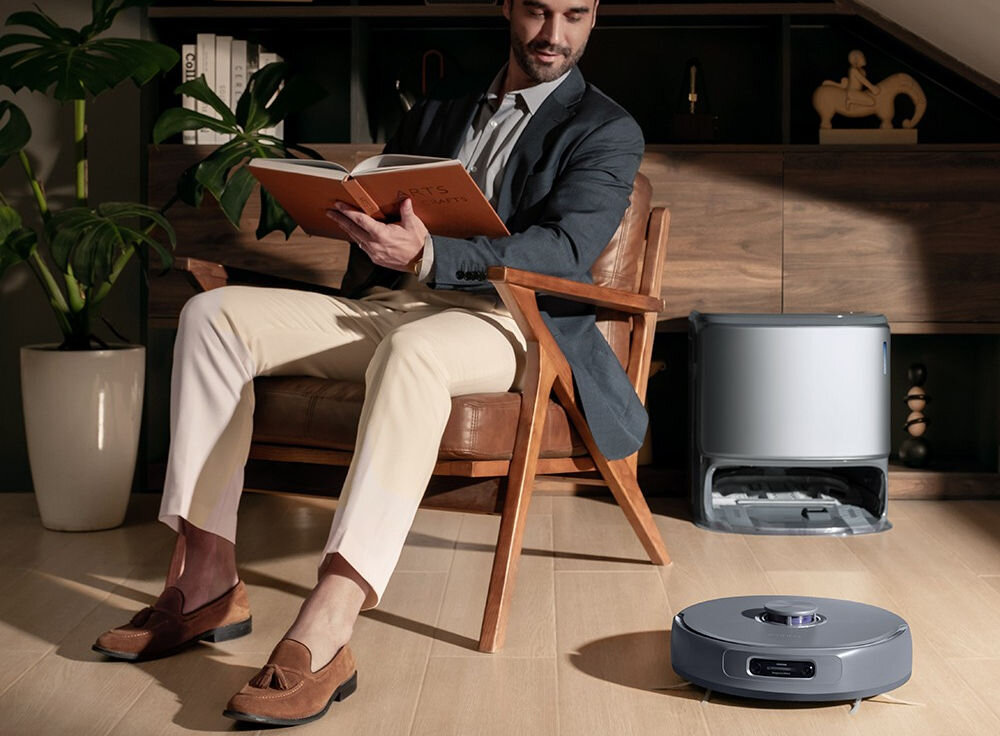 Mężczyzna siedzi na krześle i czyta książkę, obok niego na podłodze znajduje się szary robot sprzątający. W tle widać drewnianą szafkę i roślinę w doniczce.