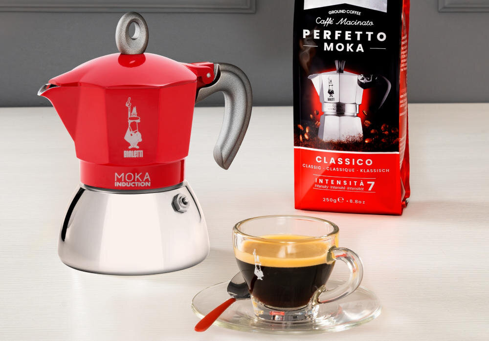 Na obrazie znajduje się czerwona kawiarka z napisem \'Bialetti Moka Induction\' oraz opakowanie kawy \'Perfetto Moka Classico Intensita 7\'. Obok stoi filiżanka z espresso na spodku z łyżeczką.