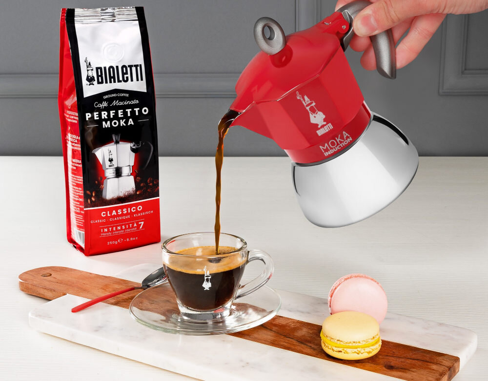Czerwona kawiarka nalewa kawę do szklanej filiżanki na drewnianej desce z dwoma makaronikami. Obok znajduje się opakowanie kawy z napisem \'Bialetti Perfetto Moka Classico Intensita 7\'.