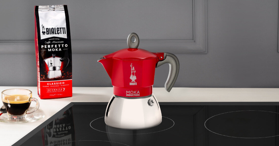 Na blacie kuchennym stoi kawiarka z czerwonym górnym elementem i srebrnym dolnym, obok niej znajduje się opakowanie kawy z napisem \'Bialetti Perfetto Moka Classico\'. Na blacie widoczna jest również filiżanka z kawą.