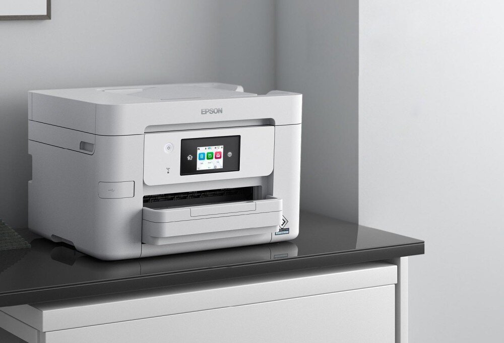 Urządzenie wielofunkcyjne EPSON WorkForce Pro WF-M4619DWF Drukarka stojąca na ciemnym blacie, ustawiona w rogu jasnego pomieszczenia, widoczny kolorowy ekran dotykowy i zamknięta taca wyjściowa, opis