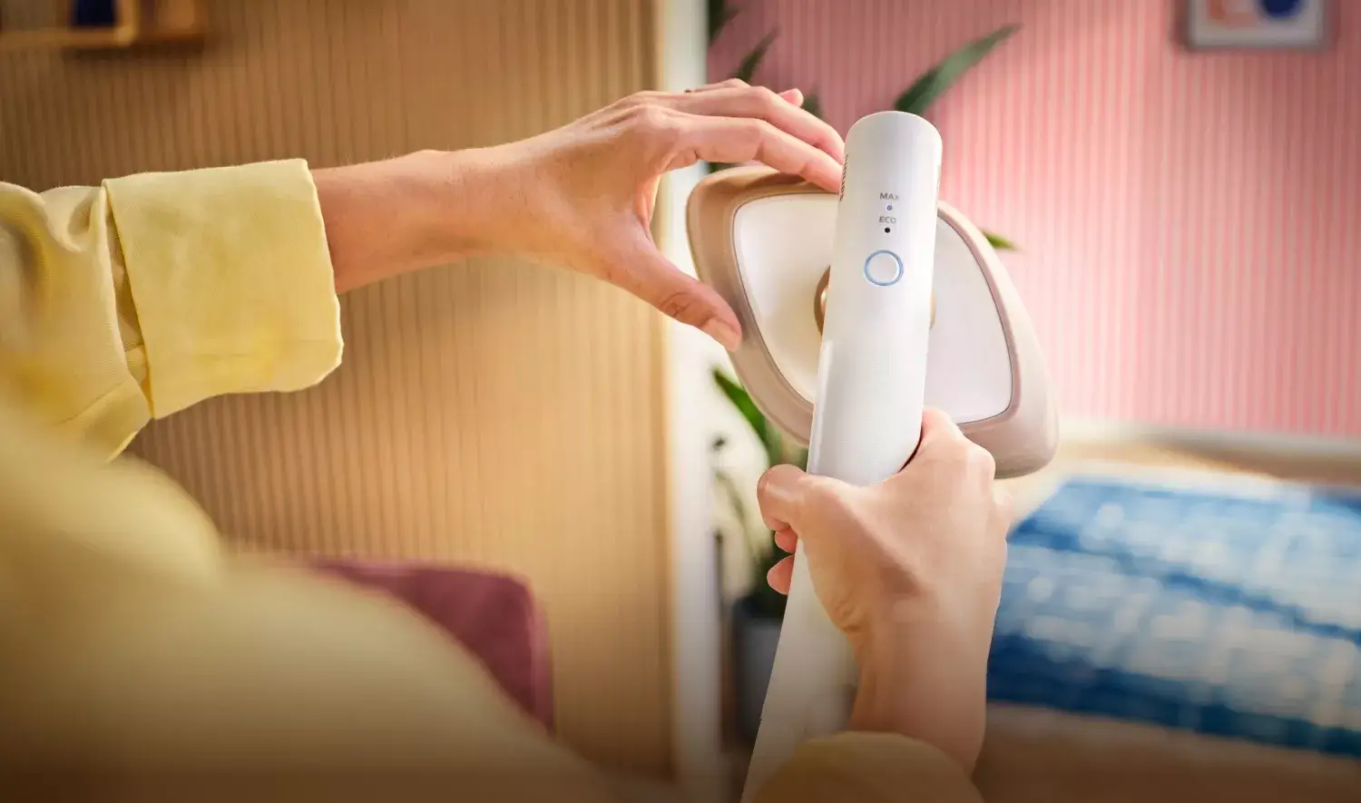 Osoba trzyma parownicę do ubrań z widocznym przyciskiem i oznaczeniami \'MAX\' oraz \'ECO\'. W tle znajduje się wnętrze z rośliną i ścianą w paski.