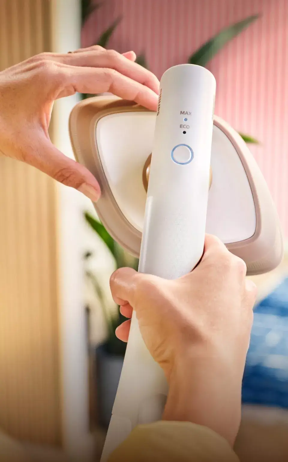 Osoba trzyma parownicę do ubrań z widocznym panelem sterowania z przyciskiem i oznaczeniami \'MAX\' oraz \'ECO\'. W tle widać rozmyte elementy wnętrza, takie jak rośliny i ściana w paski.