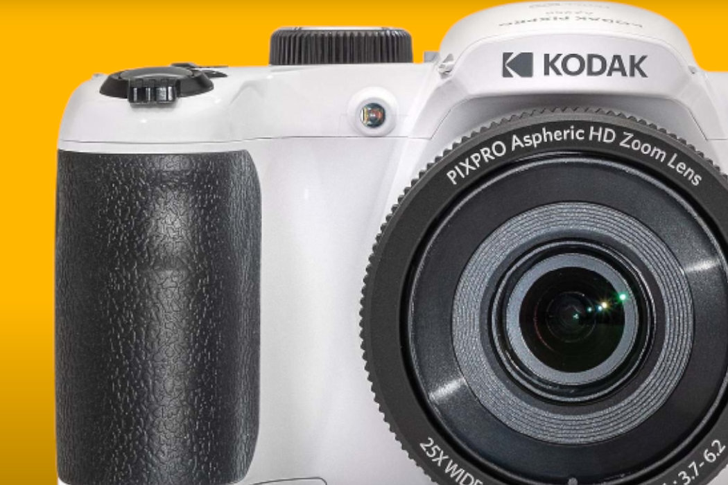 Biały aparat fotograficzny z widocznym logo KODAK oraz obiektywem oznaczonym jako PIXPRO Aspheric HD Zoom Lens. Tło jest w kolorze żółtym.