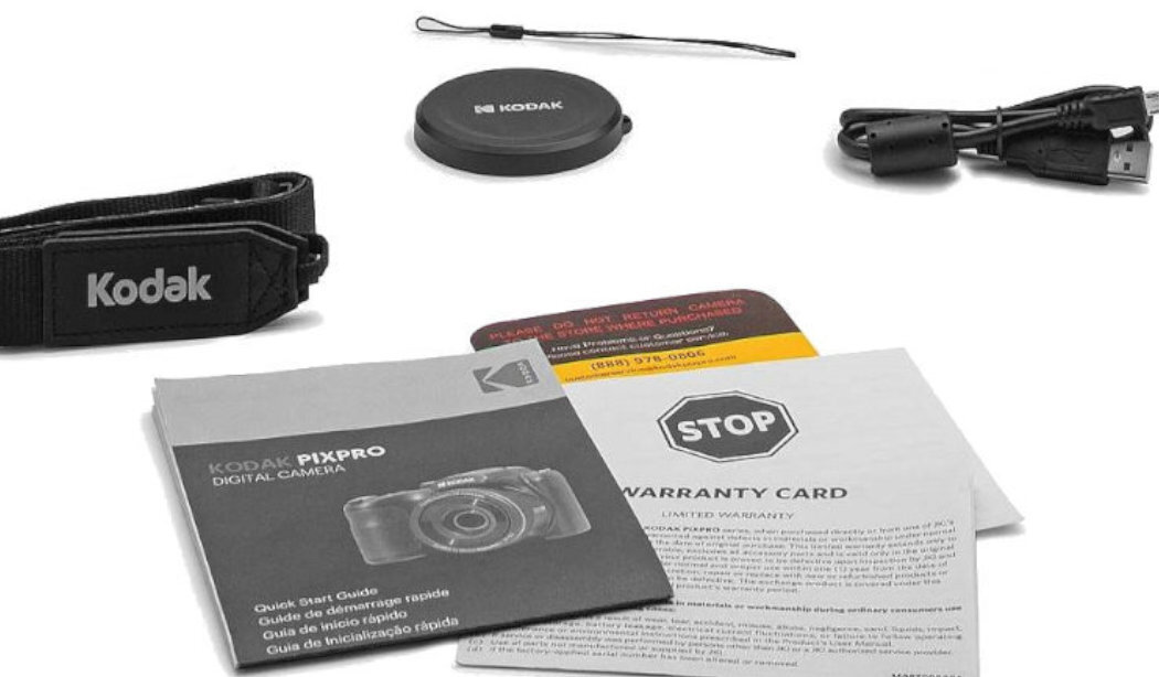 Na obrazie widoczne są akcesoria do aparatu Kodak, w tym pasek z logo Kodak, pokrywka obiektywu, kabel USB oraz dokumenty takie jak karta gwarancyjna i instrukcja obsługi. Na instrukcji widnieje napis \'KODAK PIXPRO DIGITAL CAMERA\'.