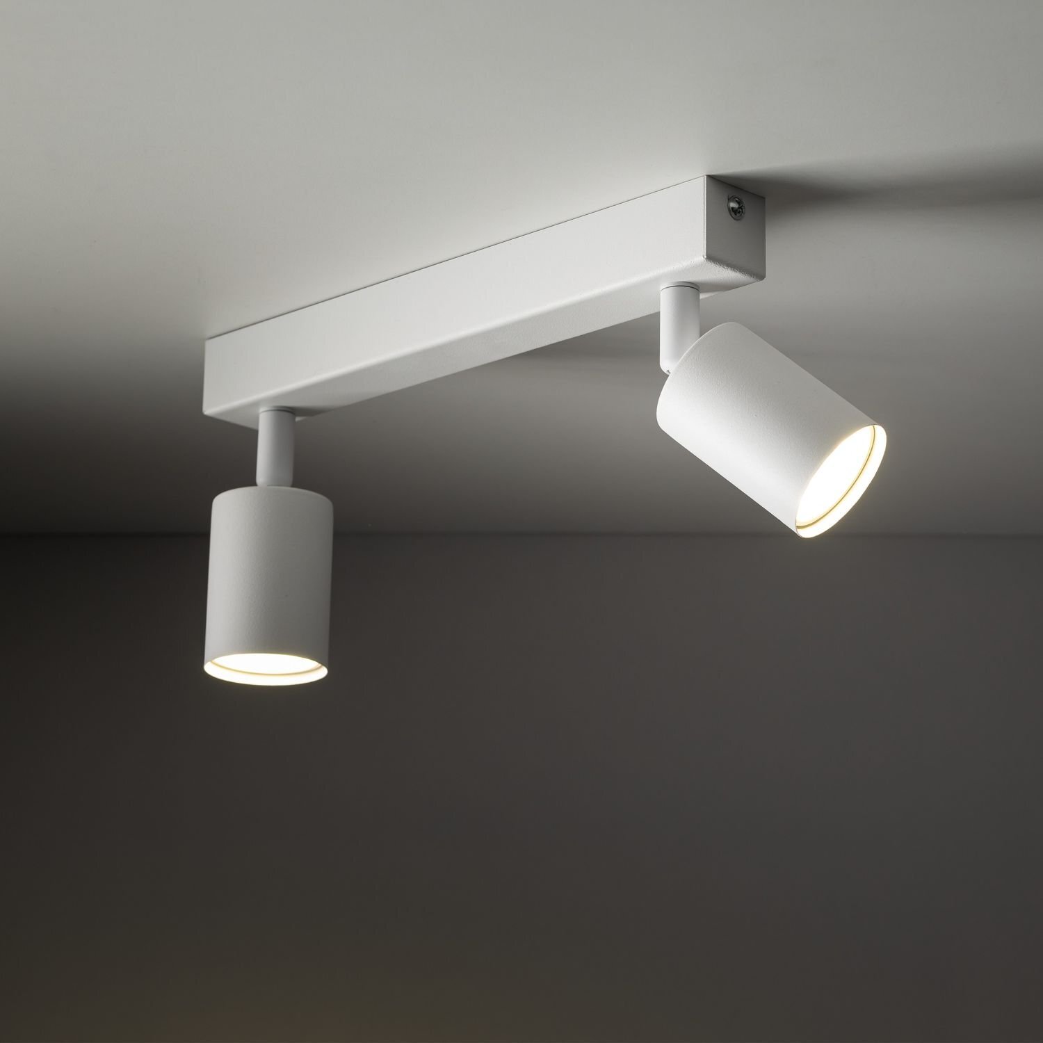 Biała lampa sufitowa z dwoma regulowanymi reflektorami zamontowana na prostokątnej podstawie. Oba reflektory są włączone, emitując ciepłe światło.