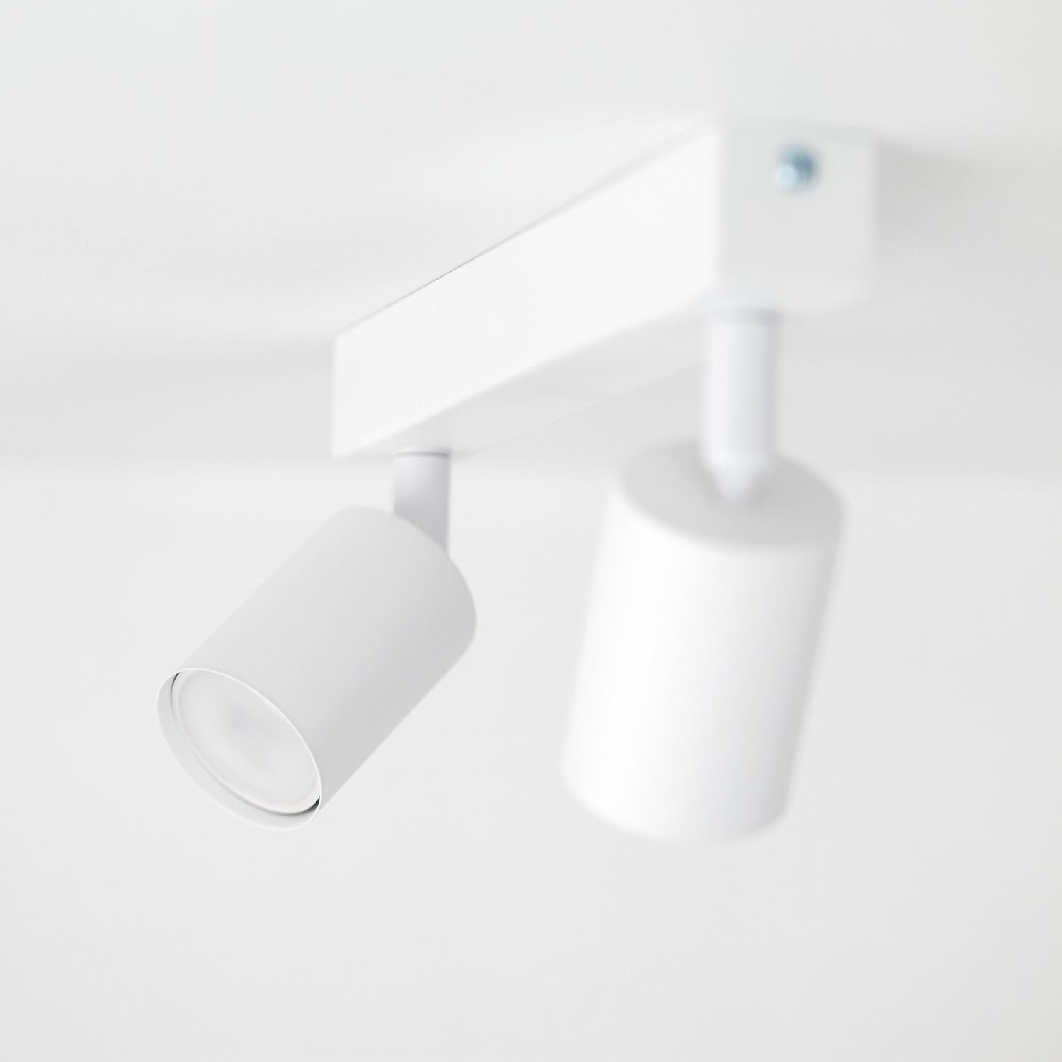 Biała lampa sufitowa z dwoma regulowanymi reflektorami w kształcie cylindrów, zamontowana na prostokątnej podstawie.