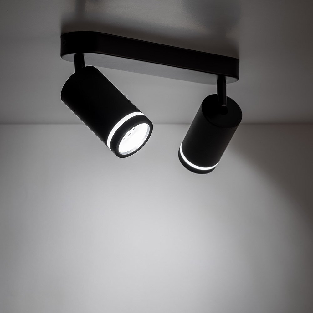 Czarna lampa sufitowa z dwoma cylindrycznymi reflektorami, które emitują światło. Reflektory są zamontowane na prostokątnej podstawie przymocowanej do sufitu.