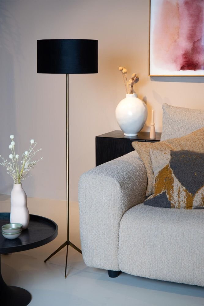 Na obrazie znajduje się elegancka lampa podłogowa z czarnym abażurem, stojąca obok beżowej sofy z poduszkami. W tle widoczny jest biały wazon na ciemnym stoliku oraz abstrakcyjny obraz na ścianie.
