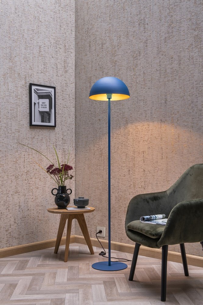 Na obrazie znajduje się niebieska lampa podłogowa z półokrągłym kloszem, stojąca obok zielonego fotela i drewnianego stolika z wazonem i książką. W tle widać ścianę z teksturą oraz obrazek w ramce.