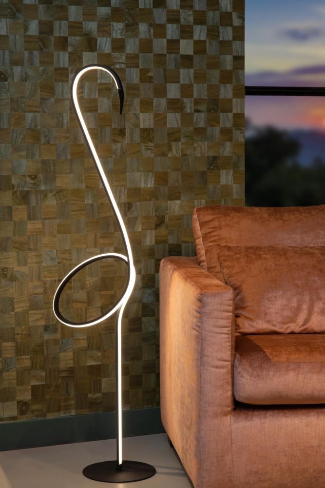 Stylowa lampa podłogowa o nowoczesnym designie, przypominająca kształtem flaminga, stoi obok brązowej sofy. W tle widoczna jest ściana z drewnianych paneli oraz okno z widokiem na zachód słońca.
