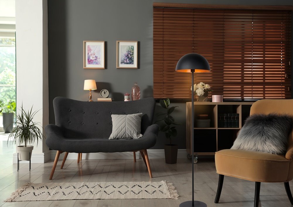 W salonie znajduje się elegancka lampa podłogowa z czarnym kloszem, obok ciemnoszarej kanapy z poduszką. W tle widać drewniane żaluzje, dwa obrazy na ścianie i regał z dekoracjami.