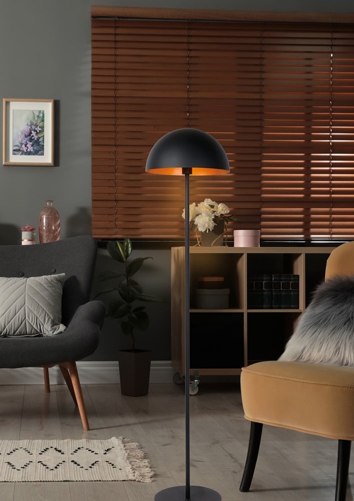 Wnętrze salonu z elegancką lampą podłogową o półkolistym kloszu, stojącą obok szarego fotela i żółtego krzesła. W tle widać drewniane żaluzje, półkę z dekoracjami oraz obraz na ścianie.