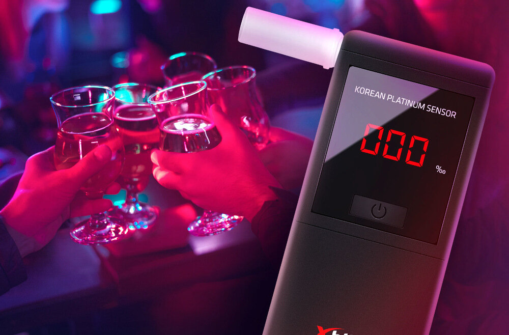 Na obrazie widoczny jest alkomat z wyświetlaczem pokazującym \'000 ‰\' oraz napis \'KOREAN PLATINUM SENSOR\'. W tle kilka osób trzymających kieliszki z napojami.