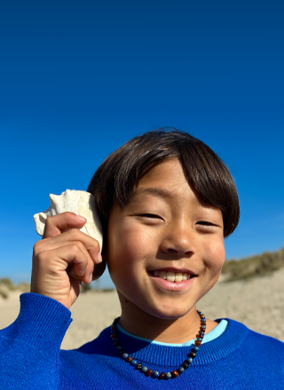 Osoba trzyma muszlę przy uchu na tle plaży i błękitnego nieba. Osoba ma na sobie niebieski sweter i naszyjnik z koralików.