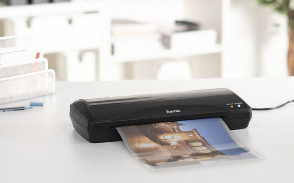 Na białym biurku znajduje się czarny laminator z logo \'hama\', z którego wychodzi zalaminowany dokument z kolorowym obrazem. W tle widoczne są rozmyte elementy biurowe.