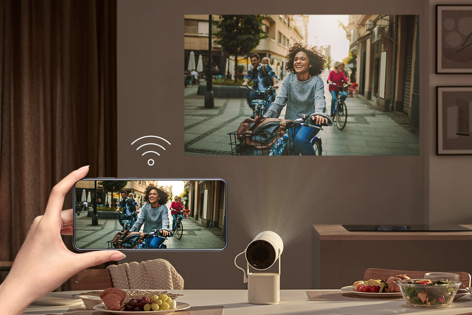 Na stole znajduje się projektor, który wyświetla obraz na ścianie przedstawiający osobę jadącą na rowerze. Obok projektora widoczny jest smartfon z tym samym obrazem, a nad nim symbol Wi-Fi.