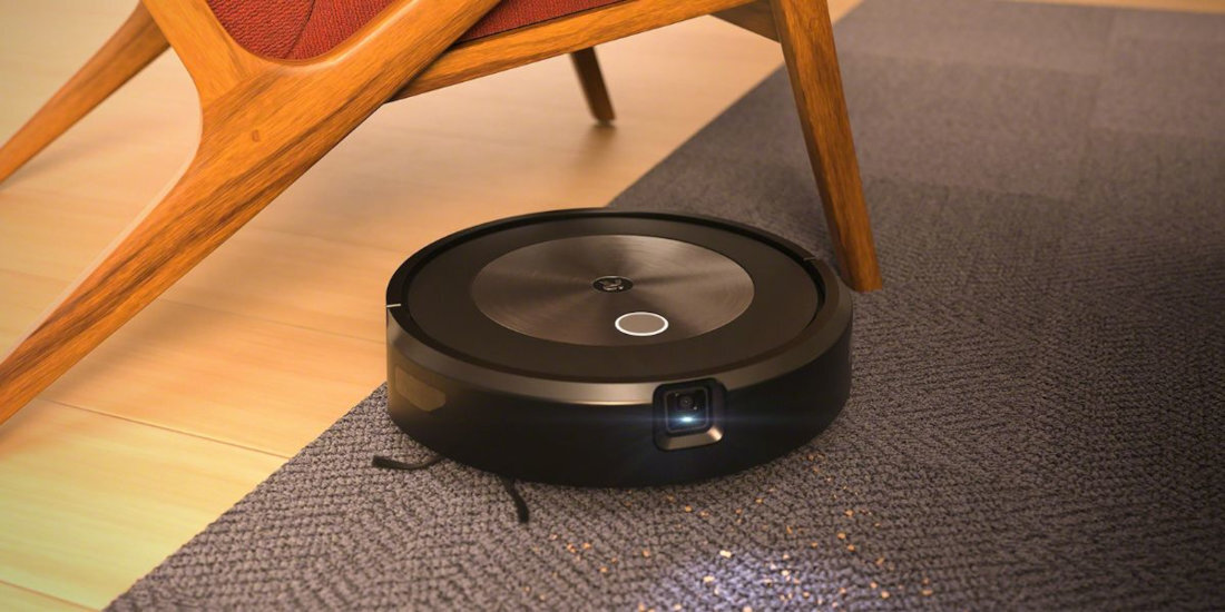 Na obrazie widoczny jest okrągły, czarny robot sprzątający na dywanie obok drewnianego mebla. Robot ma błyszczący panel na górze z centralnym przyciskiem.