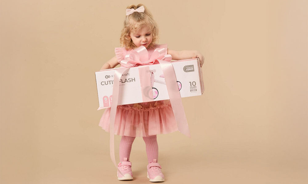 ROWEREK BIEGOWY KINDERKRAFT CUTIE FLASH PINK dziewczynka trzyma zapakowane pudełko pomysł na prezent wsparcie rozwoju dziecka doskonalenie równowagi koordynacji ruchowej ogólna sprawność fizyczna podarunek na urodziny święta Dzień Dziecka mnóstwo frajdy stymulacja zdrowego rozwoju niezapomniane chwile pełne uśmiechu