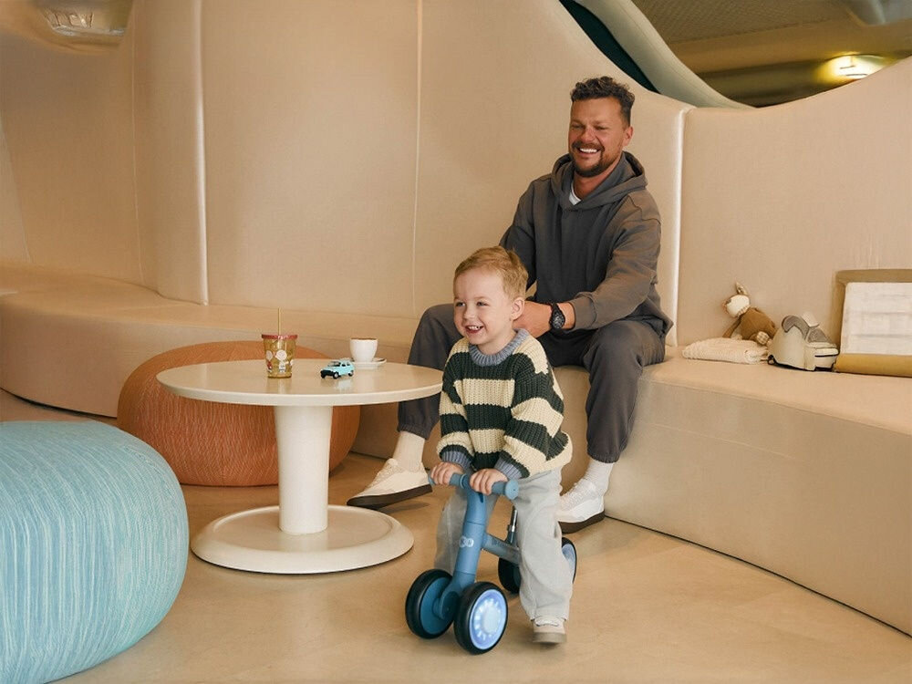 ROWEREK BIEGOWY KINDERKRAFT CUTIE FLASH PINK maluch siedzi na niebieskim rowerku obok siedzi mężczyzna dla małych miłośników aktywności kompaktowe wymiary (53 × 22,5 × 36,5 cm) lekka konstrukcja (2,1 kg) łatwy w przenoszeniu i przechowywaniu regulowane siedzisko (27–30 cm) wytrzymałe koła o średnicy 15 cm wsparcie motoryki i równowagi od 1. roku życia nauka koordynacji i samodzielnej jazdy radość z ruchu odkrywanie świata