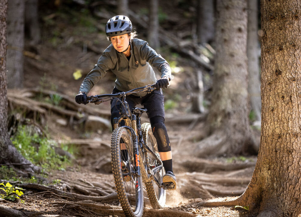 Osoba na rowerze górskim jedzie po leśnej ścieżce, nosząc czarny matowy kask rowerowy. W tle widać drzewa i korzenie na ziemi.
