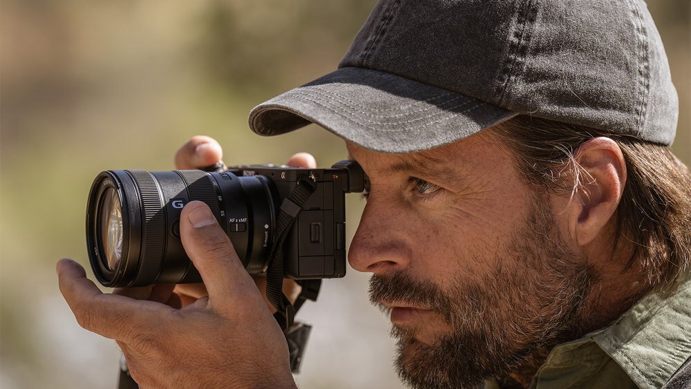 Osoba trzyma aparat bezlusterkowy z obiektywem, patrząc przez wizjer. Aparat jest czarny, a obiektyw ma oznaczenie \'G\'.