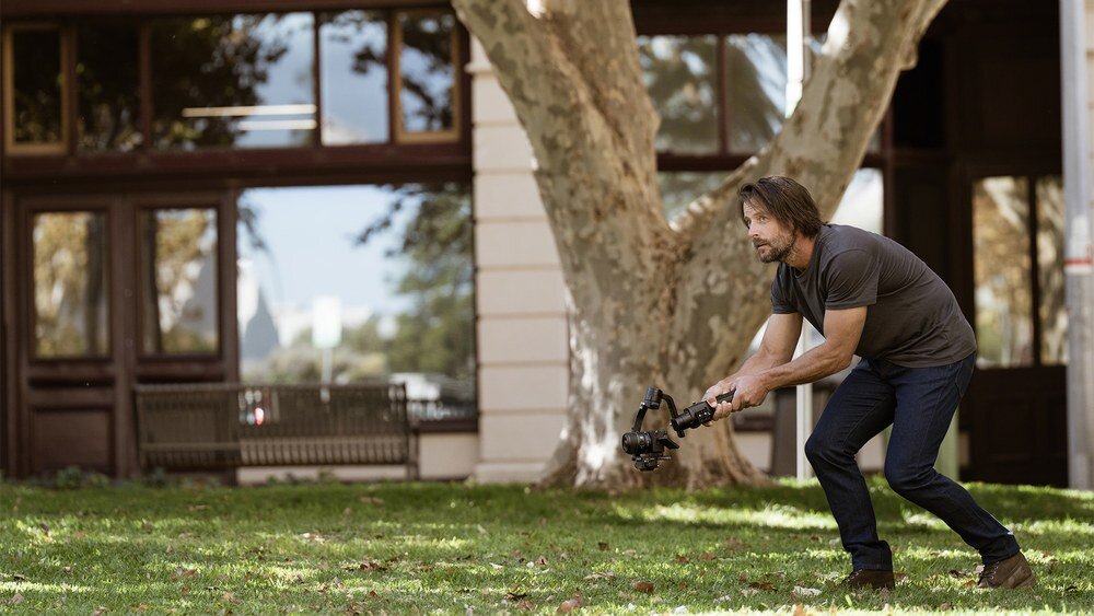 Osoba trzymająca aparat na stabilizatorze, stojąca na trawie przed budynkiem z dużymi oknami i drzewem.