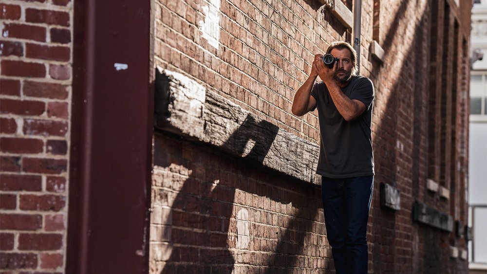 Osoba stoi przy ceglanej ścianie i trzyma aparat fotograficzny przy twarzy, jakby robiła zdjęcie. Ściana jest oświetlona światłem słonecznym, tworząc wyraźne cienie.