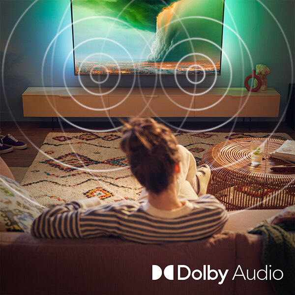 Osoba siedzi na kanapie przed telewizorem, na którym wyświetlany jest obraz z zielonymi i pomarańczowymi tonami. Na dole obrazu widnieje logo \'Dolby Audio\'.