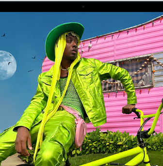 Osoba w jaskrawozielonym stroju siedzi na rowerze przed różowym budynkiem. W tle widoczny jest księżyc i kilka ptaków na niebie.