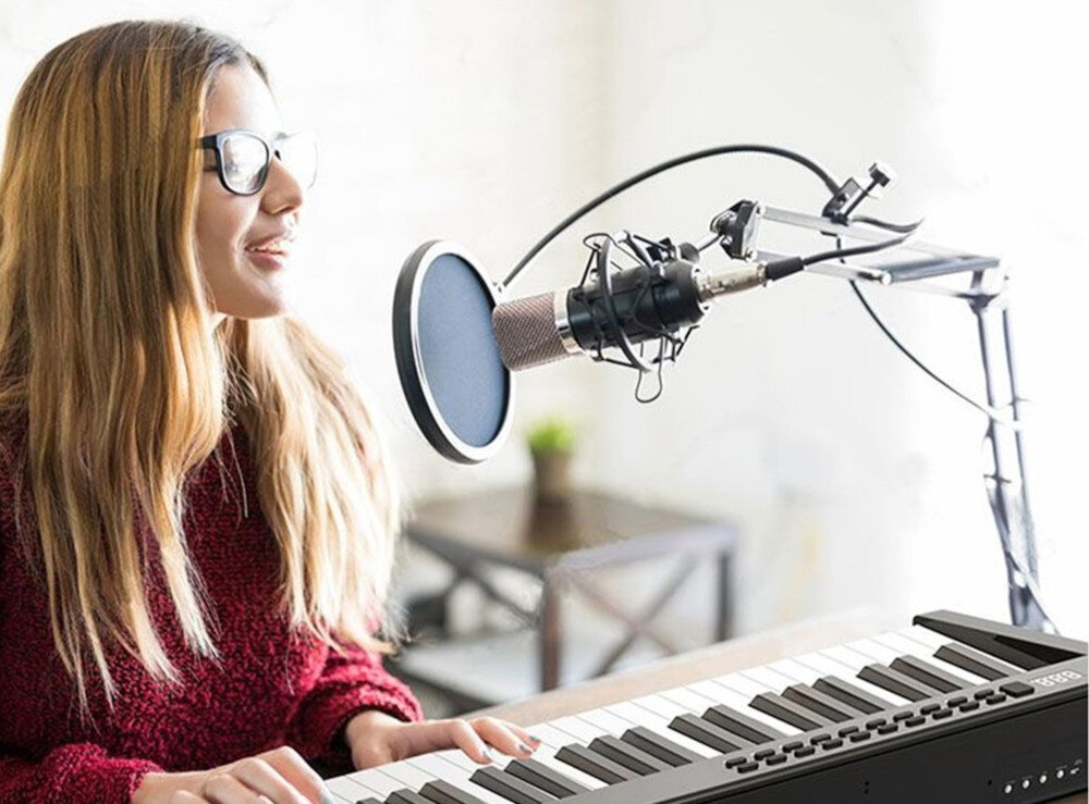 Osoba siedzi przy cyfrowym pianinie i śpiewa do mikrofonu na statywie. W tle widać rozmyty fragment wnętrza z rośliną na stoliku.