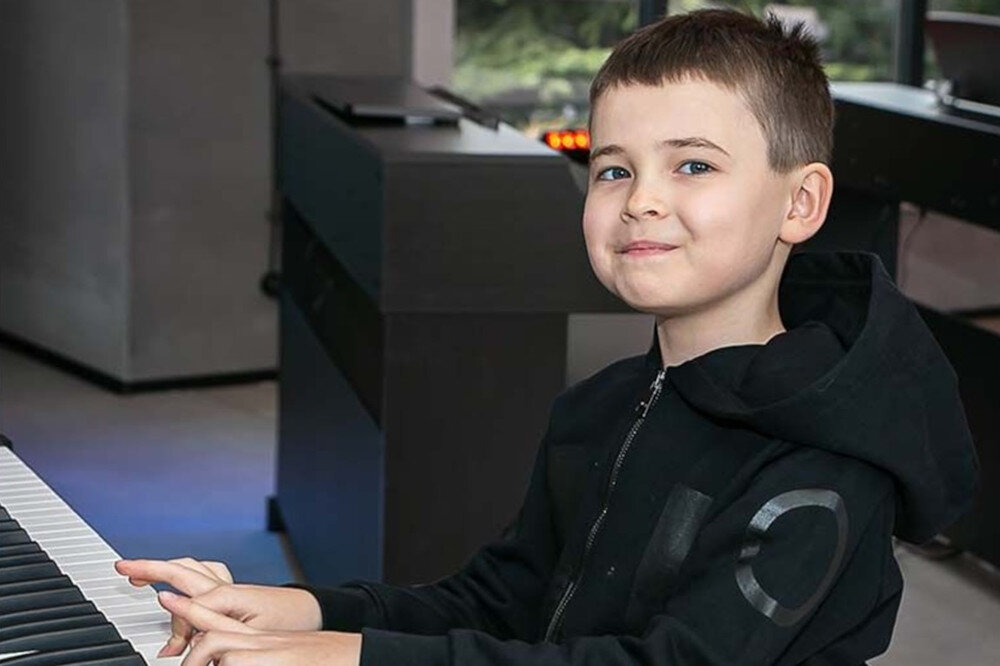 Dziecko siedzi przy pianinie cyfrowym, ubrane w ciemną bluzę z kapturem. W tle widoczne są elementy wnętrza z oknem.
