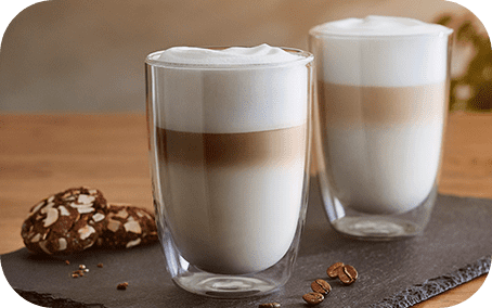 Na zdjęciu znajdują się dwa szklane kubki z kawą latte, ustawione na ciemnej tacy. Obok leżą trzy ziarna kawy i dwa ciastka.