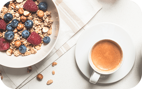 Na obrazie widoczna jest filiżanka z kawą oraz miska z musli, owocami i orzechami na jasnym tle. Obok znajduje się biały obrus w paski.