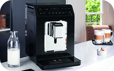 Czarny ekspres do kawy z widocznym logo KRUPS stoi na blacie kuchennym. Obok znajduje się szklana butelka z mlekiem, a w tle widać filiżanki z kawą na tacy.