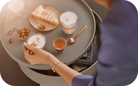 Na okrągłym stoliku znajdują się filiżanki z kawą, talerzyk z ciastem i orzechami oraz złota łyżeczka. Osoba w niebieskiej koszuli sięga po filiżankę.