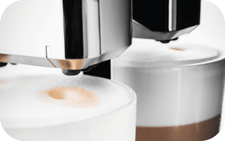 Zbliżenie na dwa kubki z mleczną pianką, pod którymi znajduje się dysza ekspresu do kawy. Kubki mają warstwową strukturę napoju z widoczną pianką na górze.