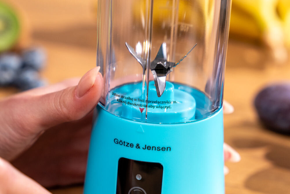 Niebieski blender personalny marki Götze & Jensen z widocznym ostrzem wewnątrz przezroczystego pojemnika. Na urządzeniu znajduje się tekst: \'Ustaw strzałkę w linii przełącznika i naciśnij dwukrotnie aby włączyć\'.