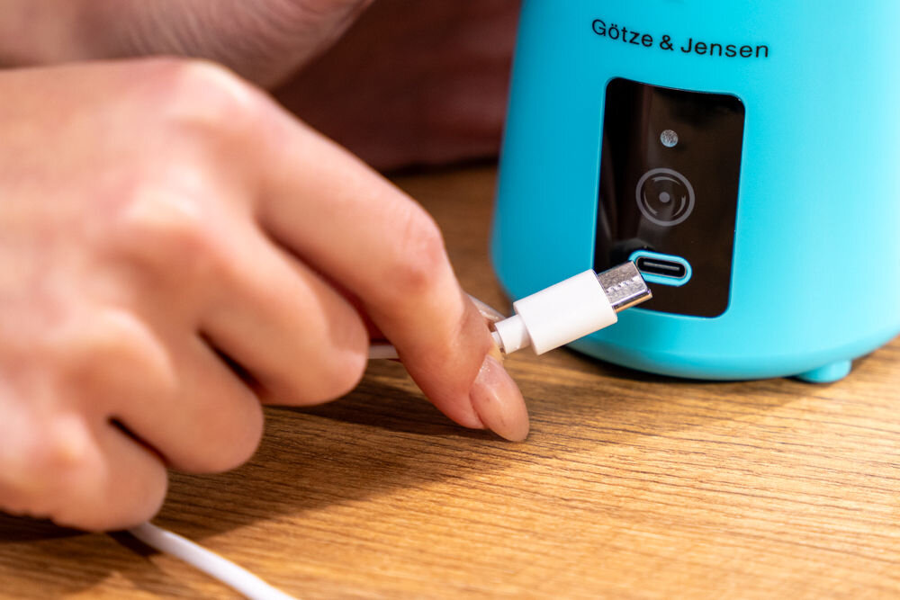 Osoba podłącza kabel USB do niebieskiego urządzenia z napisem \'Götze & Jensen\'.