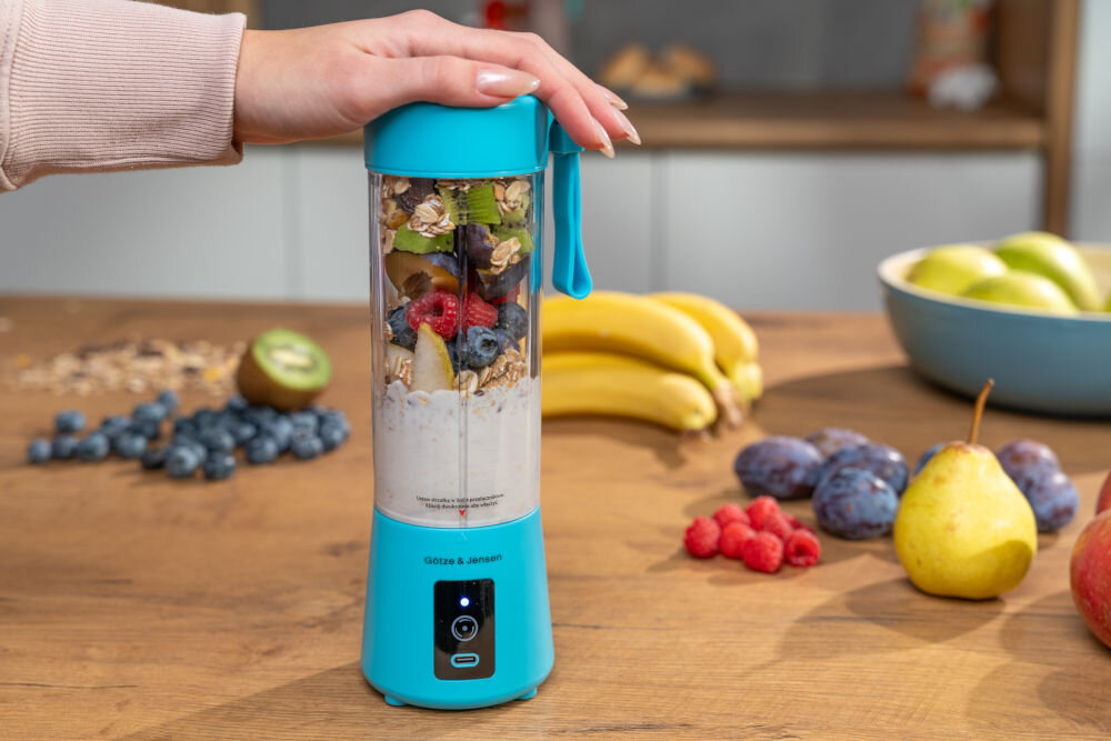 Niebieski blender personalny GÖTZE & JENSEN z owocami i płatkami w środku, trzymany przez osobę. W tle na blacie znajdują się owoce, takie jak banany, kiwi, maliny, borówki, śliwki i gruszka.