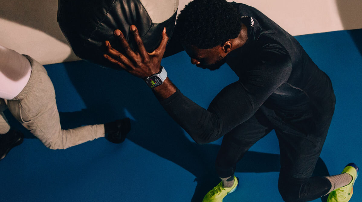 Osoba w ciemnym stroju sportowym trzyma dużą piłkę, na nadgarstku ma zegarek z jasnym paskiem. W tle widać niebieską matę do ćwiczeń.