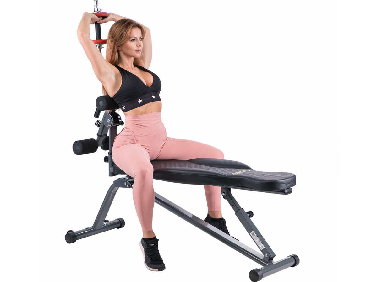 Kobieta siedzi na ławce do ćwiczeń, trzymając ręce nad głową. Ławka ma metalową konstrukcję i czarne obicie.