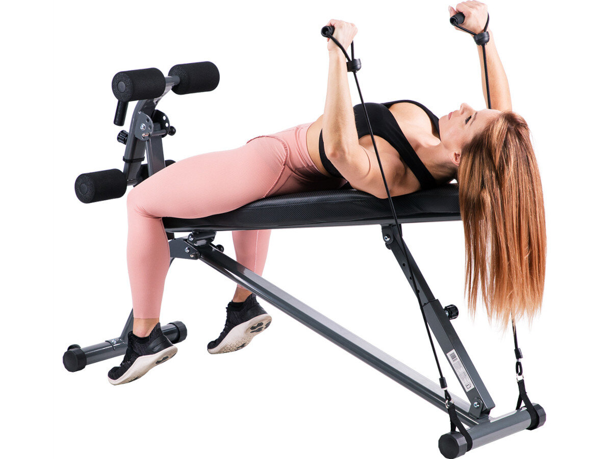 Osoba ćwiczy na ławce do ćwiczeń z gumami oporowymi, leżąc na plecach i trzymając uchwyty. Ławka ma metalową konstrukcję i czarne obicie.