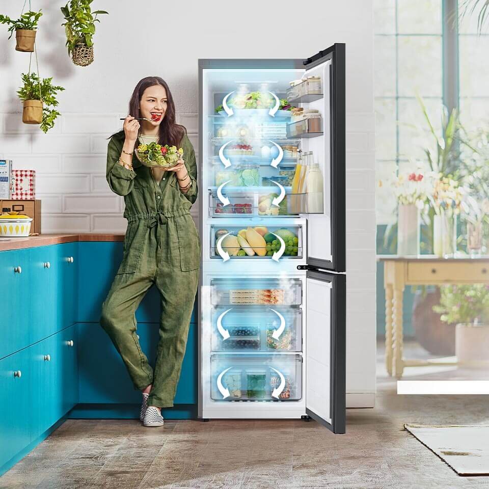 Kobieta w zielonym kombinezonie stoi obok otwartej lodówki, jedząc sałatkę. W lodówce widoczne są różne produkty spożywcze, takie jak owoce, warzywa i napoje.