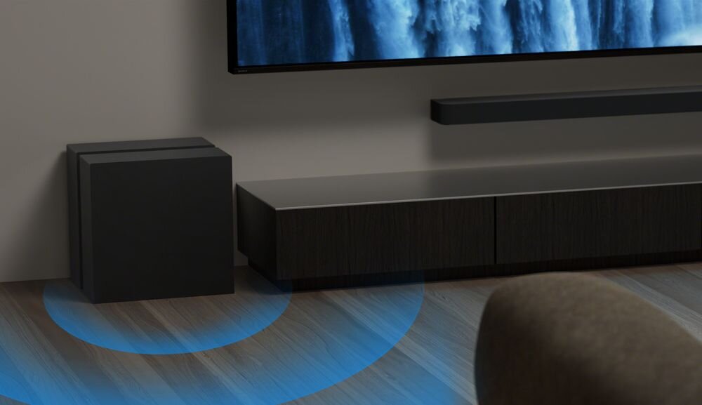 Na obrazie widać czarny subwoofer ustawiony na podłodze obok ciemnej szafki RTV. W tle znajduje się telewizor zamontowany na ścianie.