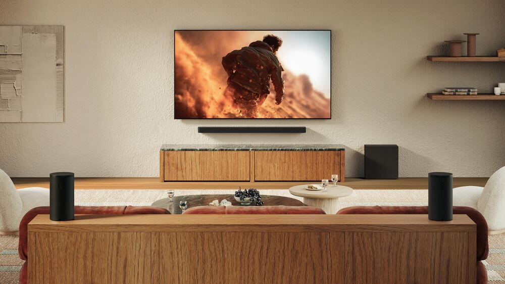 W salonie znajduje się telewizor wyświetlający scenę z filmu, poniżej zamontowany jest soundbar. Na podłodze obok szafki stoi czarny subwoofer.