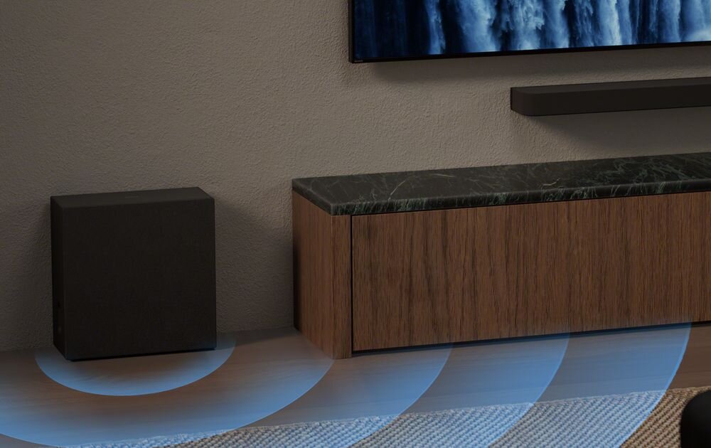 Na obrazie widać czarny subwoofer ustawiony obok drewnianej szafki z ciemnym blatem. Na podłodze widoczne są niebieskie fale dźwiękowe.