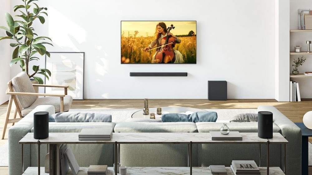 W salonie znajduje się telewizor na ścianie, na którym wyświetlany jest obraz kobiety grającej na wiolonczeli w polu. Przed telewizorem stoi szara sofa, a obok niej widoczny jest czarny subwoofer.