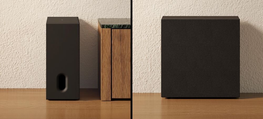 Na obrazie widoczny jest czarny subwoofer o prostokątnym kształcie, umieszczony na drewnianej powierzchni obok drewnianej szafki. Ściana w tle jest jasna i ma teksturę przypominającą tynk.